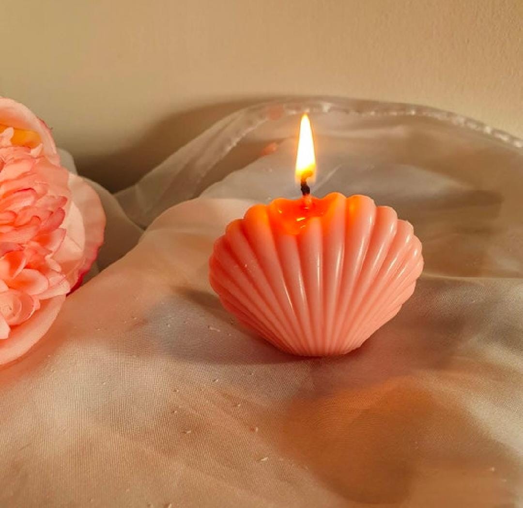 Pink shell-shaped candle with a flame on a reflective surface.
