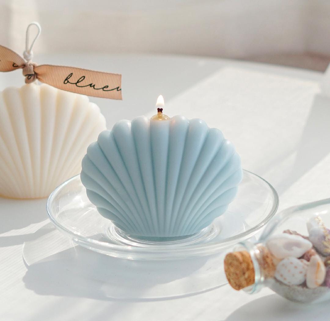 Blue shell-shaped candle on a white surface with a small bottle of seashells and a tag in the background.