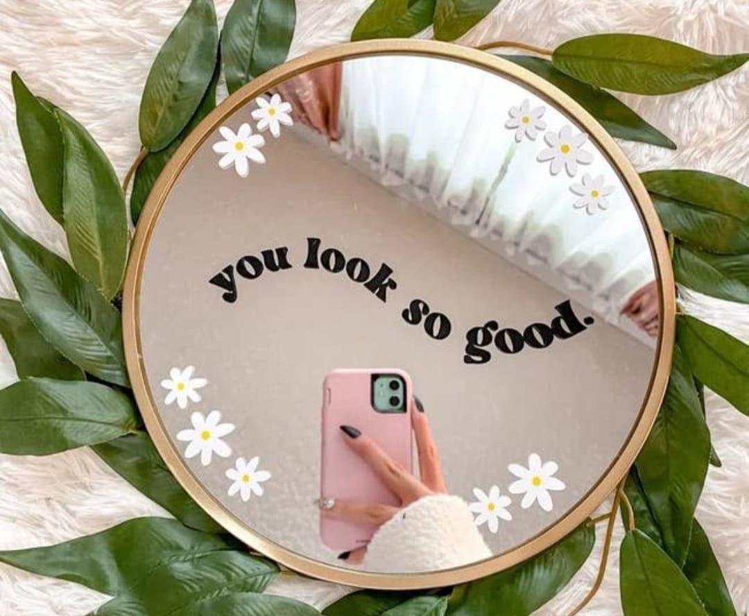 Round mirror with 'you look so good' text and floral decorations, surrounded by green leaves.