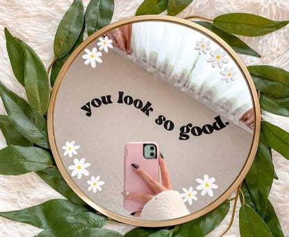 Round mirror with 'you look so good' text and floral decorations, surrounded by green leaves.